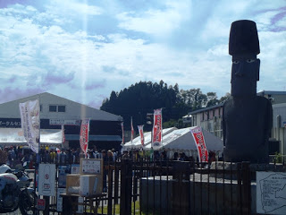 屋外に設置された大きなモアイ像の後ろで、のぼりを立てた白いテントが立ち、多くの人々で賑わう福興市が開催されている様子の写真