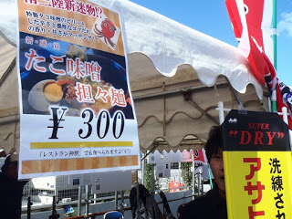 白いテントの柱に「たこ味噌担々麺 ¥300」と書かれた写真付きポスターが掲げられ、その横に「アサヒスーパードライ」の黄色い看板が立つ屋台の様子を撮影した写真