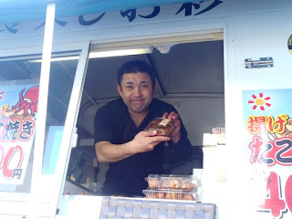 キッチンカーの窓から黒い服を着た男性が笑顔でたこ焼きのパックを差し出している様子の写真