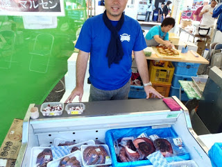青いシャツを着た男性が、冷蔵ケースに並べられた生や加工品のタコを販売する屋台に立っている様子の写真
