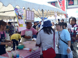 白いテントの下、赤いチェック柄のテーブルクロスの屋台で男性店員が調理を行い、来場者が列を作っている賑やかなイベント会場の様子を撮影した写真