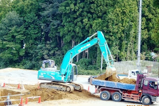 青いショベルカーが土をすくい上げて赤いダンプトラックに積み込んでいる様子を写した土の運搬作業中の写真