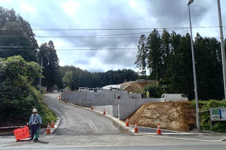 舗装された新しい道路の入口にカラーコーンと案内板が設置され、奥で工事車両が出入りしている造成工事の入口部分を写した写真