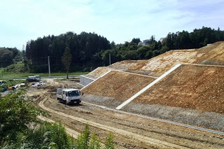 整地が進み傾斜地にコンクリートの擁壁が施工され、下の工事用道路をトラックが走行している様子の写真
