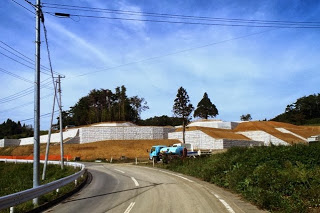 舗装された道路のカーブの先で、造成工事が進み、斜面に白い擁壁が設置されて整地が進んでいる様子の写真