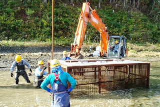 周囲にはヘルメットをかぶった作業員たちが立って見守るなか、オレンジ色のショベルカーが川の中で金属製の檻を作っている、やな場の設置作業の写真