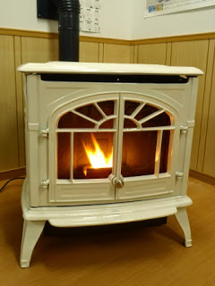 室内の木目調の壁の前に設置された白いクラシックなデザインの薪ストーブの中で、オレンジ色の炎が静かに燃えている様子が写っている写真