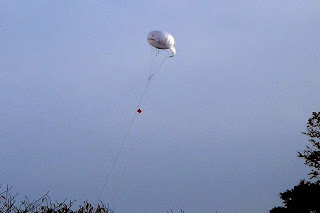 灰色の空に白い臨時用無線中継システムが細いワイヤーでつながれて飛んでいる写真