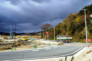 重機やクレーン車が工事をし地面はまだ舗装されておらず土がむき出しで、資材や土砂が積まれている工事現場の側をトラックが走っている写真