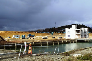 たまりをパイプの柵で囲んで設置され、白い建物と造成中の茶色い土の盛り土が広がり、重機や車両が並び、空には黒い雲が広がる造成地の写真