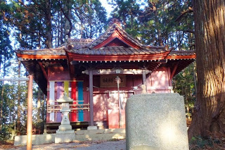 赤い屋根と柱が特徴的な日本の神社が木々に囲まれて建っており、手前には石灯籠が置かれている写真