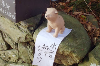 石の上に小さな犬の置物が置かれ「ボクを拾ってください」と書かれた布が垂れ下がっている写真