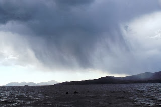 暗い雨雲が空一面に広が、遠くの山と海上に雨がふり、風で波が立ち荒れている様子の写真