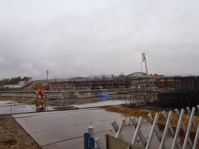 右奥にクレーンが伸び、鉄筋が組まれている雨で濡れた地面に足場が設置された、コンクリート基礎工事が進行中の写真