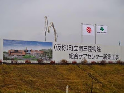 クレーンが作業を行っている手前に「仮称 町立南三陸病院 総合ケアセンター新築工事」と書かれた看板が立ち、建物完成予想図と工事用の旗が並ぶ建設現場の案内看板の写真