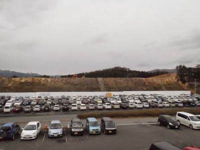 曇り空の下、斜面に囲まれた造成地の前に白いフェンスが伸びており、その前には数十台の車が密集して駐車された造成地の写真