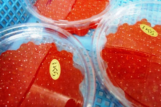 透明のプラスチック容器に入った鮮やかな赤いイクラが、青いかごに並べられて販売されている写真