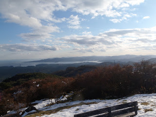 雪が残った草原に木製ベンチがあり、その奥に広がる赤茶けた木と山々、遠くに海が広がり柔らかい光に照らされた写真