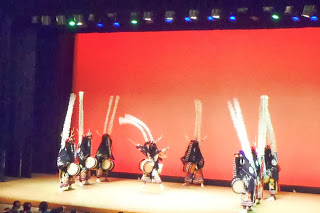 赤い背景の舞台上で、黒い装束と鹿角を模した長い白い竹飾り（ササラ）を背負った踊り手たちが太鼓を叩きながら並び、一人の踊り手が大きく身体を反らせて跳躍している、行山流水戸辺鹿子躍の写真