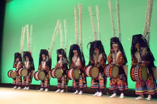 緑色の背景の舞台に、顔を白布で覆い、鹿角を模した長い白い竹飾り（ササラ）を背負い、黒頭巾と派手な柄の袴、腰太鼓を身に着けた八人の踊り手がが横一列に整列して並んでいる様子の写真