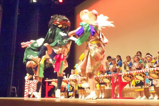 赤い背景の舞台で、緑色の布を被った大きな獅子頭を持つ演者と、笠に花飾りをつけたカラフルな衣装の踊り手が向かい合い、激しい動きで舞いながら演じており、背後には揃いの法被姿で太鼓と笛を演奏する子どもたちが並んでいる、子どもたちによる獅子舞と踊りの写真