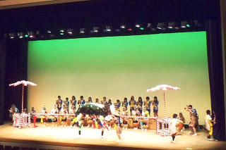 緑の背景の舞台上に大勢の子どもたちが並び前列は太鼓、後列は笛を演奏しており、その前で獅子頭をかぶった演者と、色鮮やかな衣装に菅笠を被った踊り手たちが、舞台いっぱいに広がって踊っている、子どもによる郷土芸能発表の写真