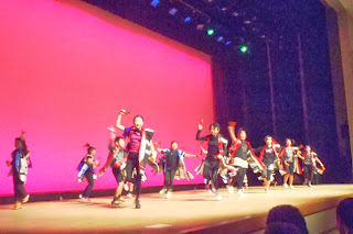 赤い背景の舞台上で、揃いの法被のような衣装を着た生徒たちが手を高く上げたりジャンプしたりしながら、元気よく一斉に踊っているよさこいの踊りを披露している様子の写真