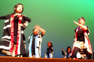 緑の照明が当たるステージ上で、揃いの黒と白の縞模様の法被を身に着けた生徒たちが、腕を大きく動かしてよさこいの踊りを披露している写真