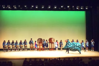 緑色の照明が当たる舞台上で、青い法被を着た子どもたちが和太鼓や笛を演奏しており、右側では緑の模様がついた大きな獅子舞が左右に動きながら演技をしている演目の様子の写真