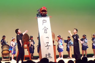 獅子舞が高い位置から「戸倉っ子魂」と書かれた垂れ幕を掲げており、両脇では黒い服を着た二人の人物が、獅子舞を紹介する振り付けで両手を広げている演出の一場面を写した写真