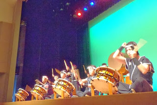 ステージ手前から見上げる角度で、黒い衣装とはちまきを身につけた太鼓奏者たちが真剣な表情で小太鼓を打っている迫力ある演奏中の写真