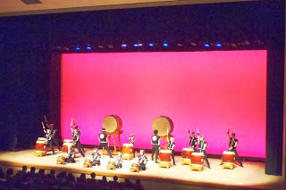 赤い照明の下、大型の和太鼓を中心に黒い衣装の奏者たちが一糸乱れぬ構えで太鼓を打つ姿が広角で捉えられた力強い演奏中の写真