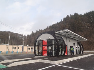 黒とグレーの丸みを帯びたデザインの建物が山のふもとに建っていて、建物の前には青い上着と反射板を着た人が立っている志津川駅の駅舎の写真