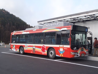 舗装され乗降場に赤いボディにキャラクターのイラストが描かれたハイブリッドバスが停車しており、乗客が次々と乗車している写真