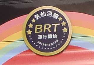 虹色のにペイントされたバスの車体に「気仙沼線BRT運行開始2012年12月22日」と書かれた金色で縁取られた黒い丸いプレートのヘッドマークが取り付けられている写真