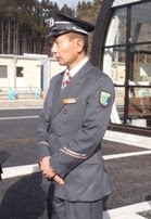 グレーの制服にネクタイと制帽を着用した男性運転手が、ガラス張りの建物の前で腕を組んで立っている写真