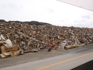 背景には曇り空と遠くの山々が見え、道路沿いにオレンジ色の三角コーンが立つ奥に、木材や金属などの建築資材が混ざった瓦礫の山が広がっている写真