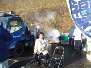 青いトラックのそばで蒸気の立ち上る鍋の前で、白い器と箸を持って椅子に座っている男性を写したイベントの様子の写真