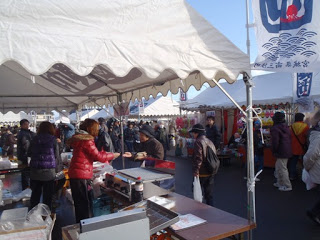 白いテントが並ぶ屋外の会場で、テーブルに商品が並べられ、買い物客が店の人とやりとりをしながら品物を手に取ったりしている様子が映された福興市の写真