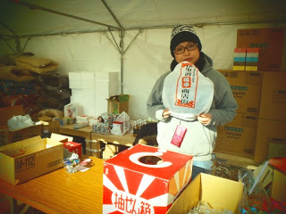 テントの中でニット帽をかぶった女性が『特賞 南三陸町商工会』と書かれた紙袋を手に持ち、景品や箱が並ぶ抽選コーナーの様子を写した写真