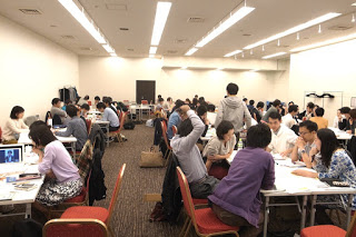 明るい照明の下、白いテーブルと赤い椅子が並ぶ広い室内で、多くの人々がグループに分かれて座り、ノートパソコンを使ったり話し合ったり書き物をしているワークショップの様子の写真