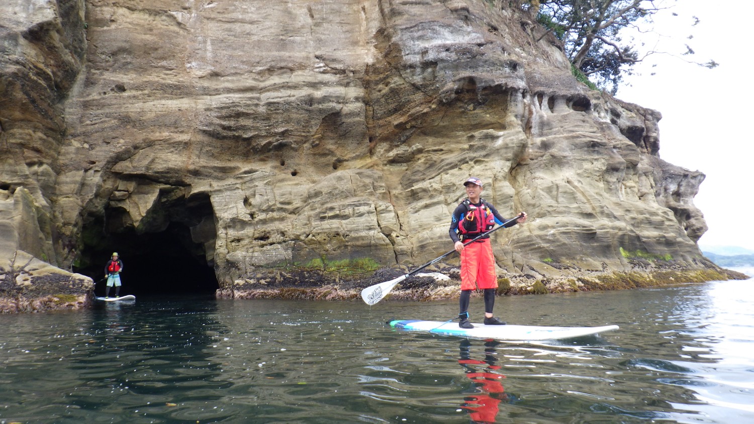 洞窟の入り口がある切り立った岩壁の前で、スタンドアップパドルボードに乗った人物がパドルを持って海の上に立っている写真