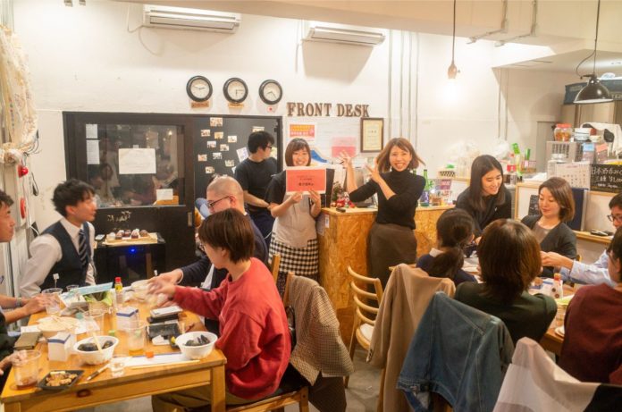 2つのテーブルでたくさんの参加者が食事を楽しむ中、「FRONT DESK」と書かれたカウンターの前に2人の女性が立ち、1人が両手でカウンターの方を示し、もう1人が赤い紙を掲げている様子の写真