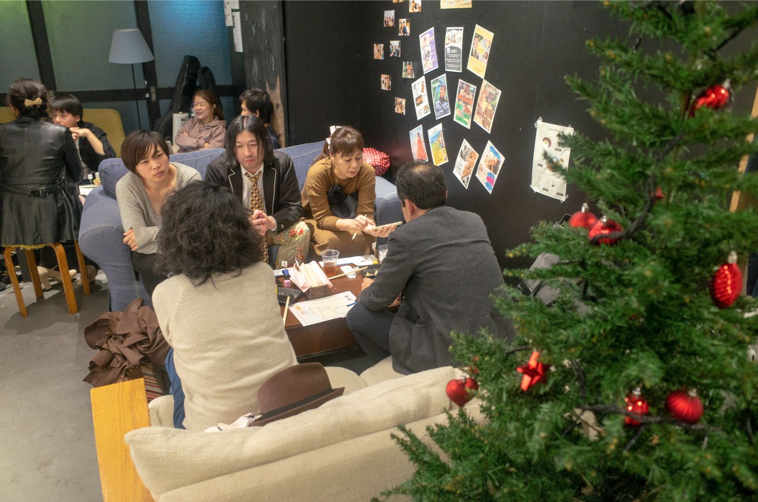 手前にクリスマスツリーがあり、黒い壁に色とりどりのチラシや写真が貼られ、テーブルを囲んで5人で食事を楽しむ様子の写真