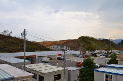 工事用フェンスの奥に造成中の盛土が広がっており、手前には仮設事務所や車両が並んでいる造成地周辺の駐車場の写真