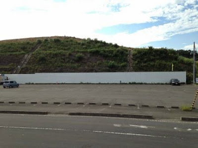 斜面に草が生えた盛土の前に白い工事用フェンスがまっすぐ設置されており、手前には広いアスファルト舗装の駐車場がある造成地の写真