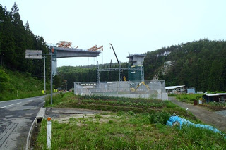 周囲には山と田畑が広がり、左側に道路が通っている山に挟まれた谷で工事が開始されたばかりの、両側の橋台が宙に浮いた状態の工事初期段階の橋の写真