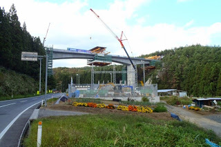 道路沿いには季節の花が植えられている奥で橋の建設が進み、クレーンや重機が橋の中央部をつなぐために稼働している、工事中期の橋の写真