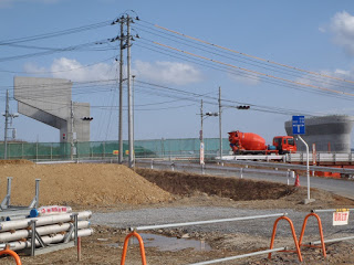 手前にガードレールと工事用資材が置かれ、奥には建設中のコンクリートの橋脚と赤いミキサー車が見える道路工事現場の写真