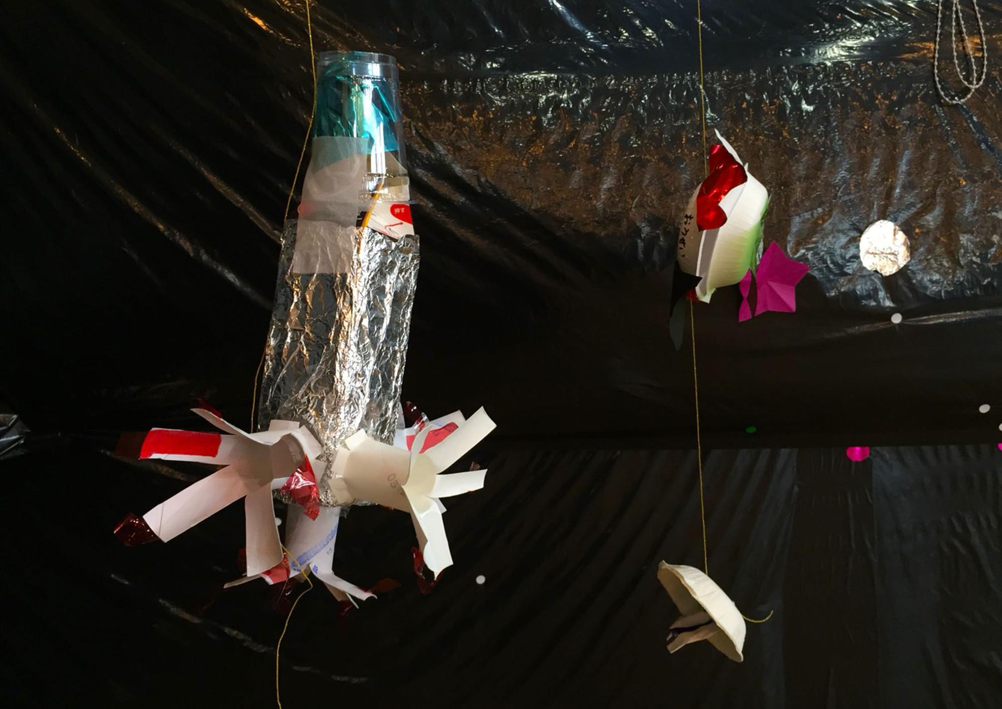 壁に真っ黒なシートが貼られ、天井から糸で吊るしたロケットやユーホーなどの作品の写真